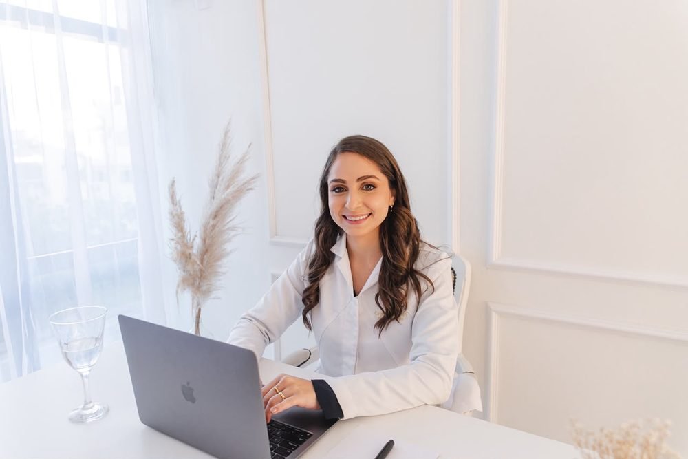 Dra. Cristina Haygert de jaleco branco, sorrindo à mesa do consultório com o notebook